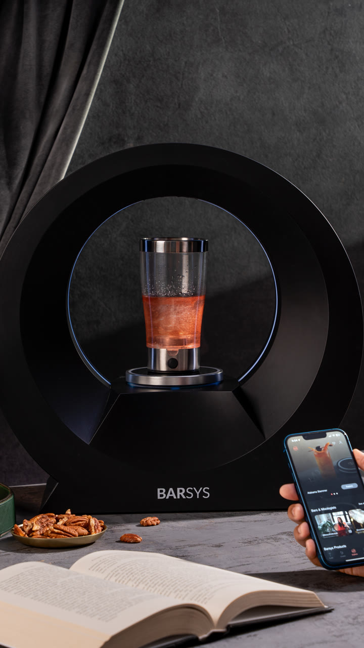 A Barsys 360 pours a red cocktail in low light beside a phone with the app.